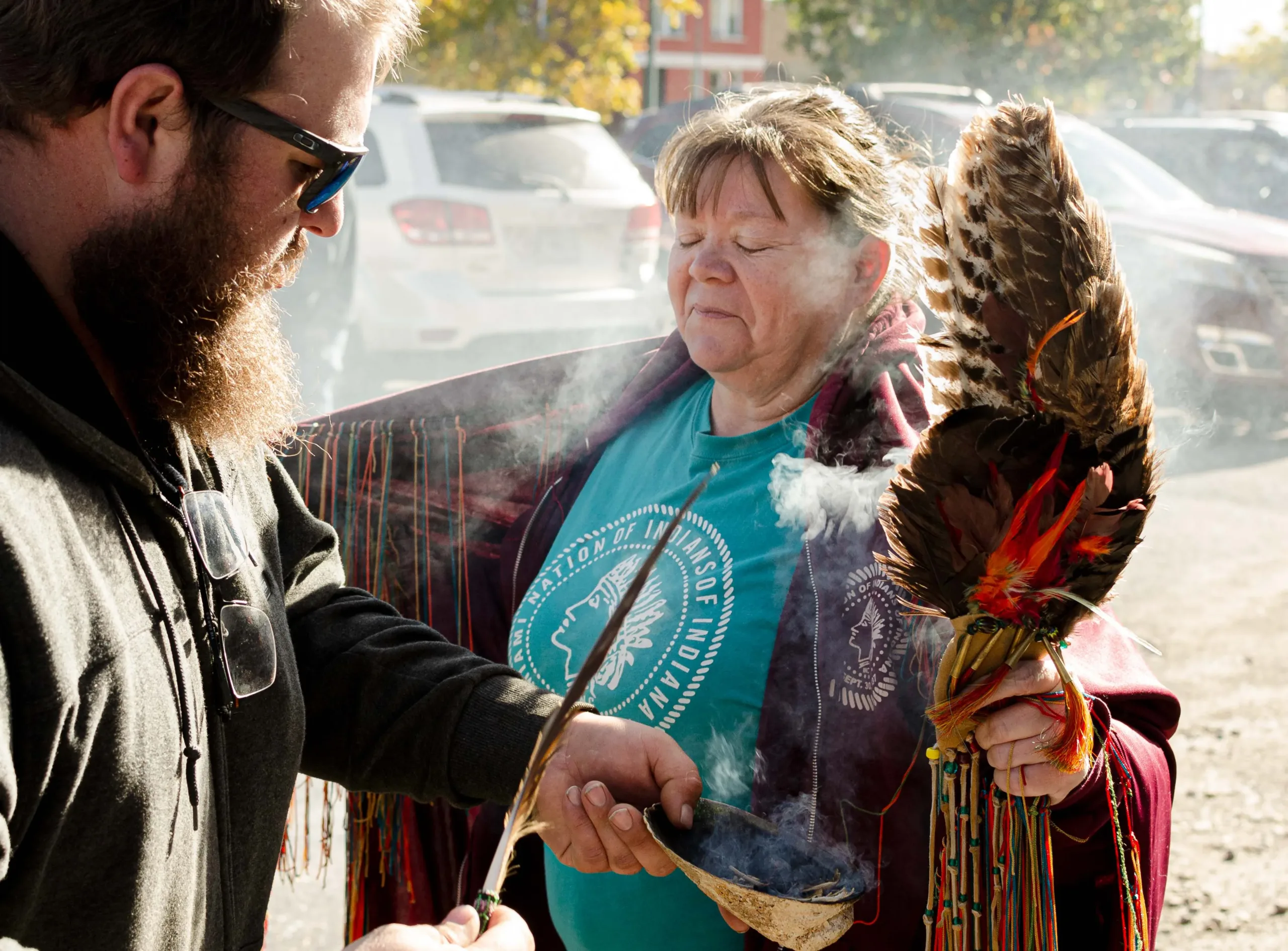 A man wearing a sweatshirt holds a feather in his right hand and a shell burning incense in his left hand. Smoke billows up and surrounds a woman wearing a maroon shaw with yellow and red tassels.