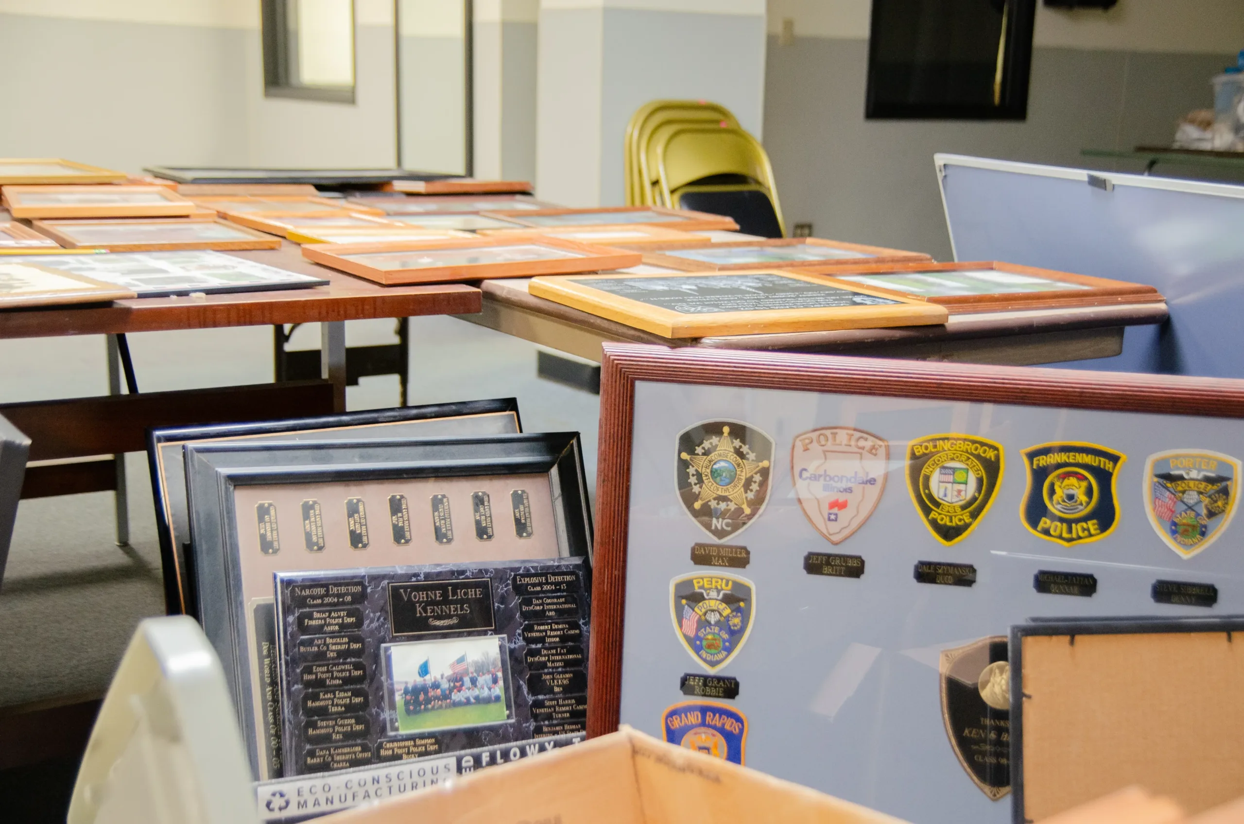 Picture frames lay on tables in the background, with a large picture frame containing police badges sits in a box in the foreground.