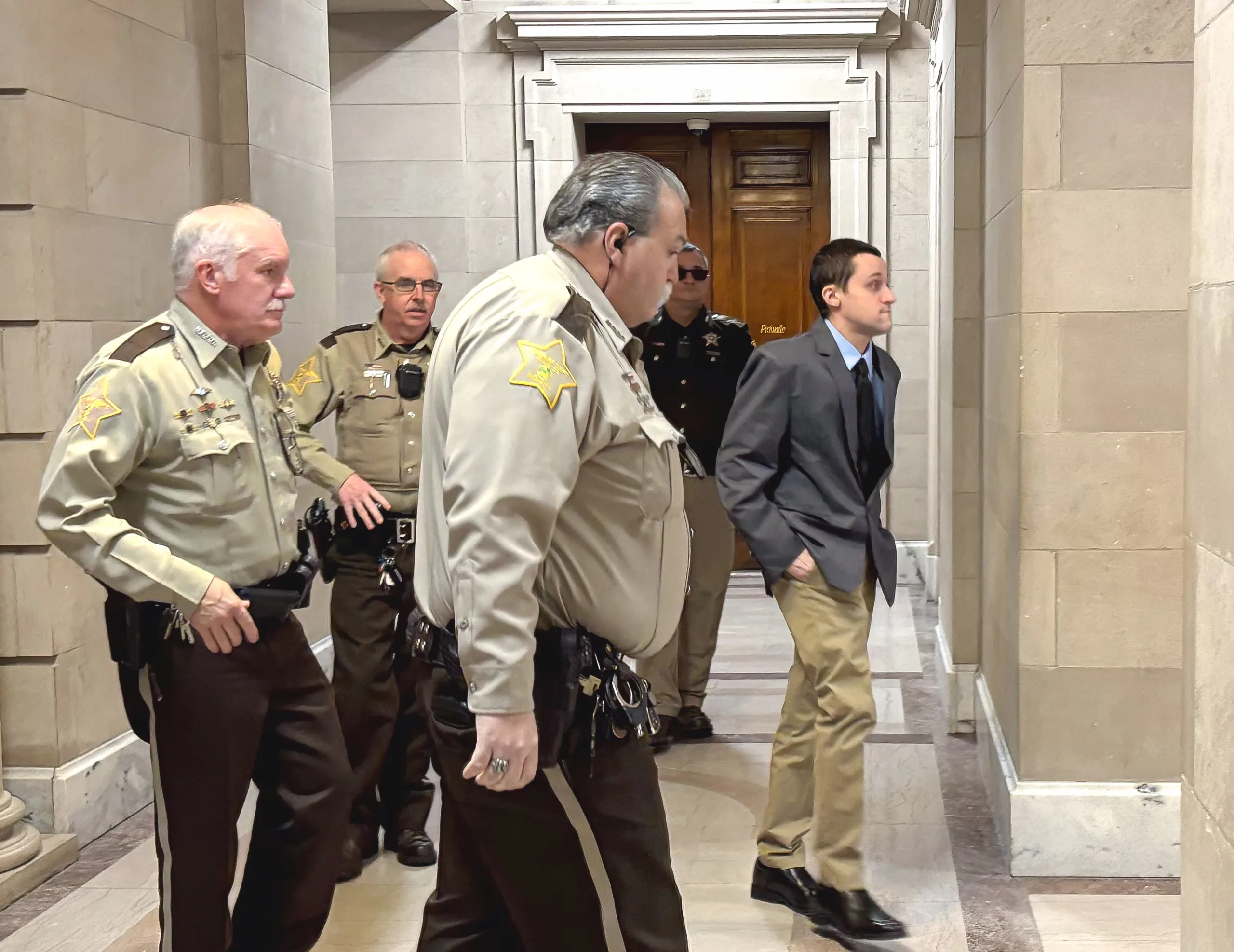 Officers wearing tan uniforms walk with Mitchell Page, dressed in a grey suit jacket and khaki pants, down a hallway with tan concrete.