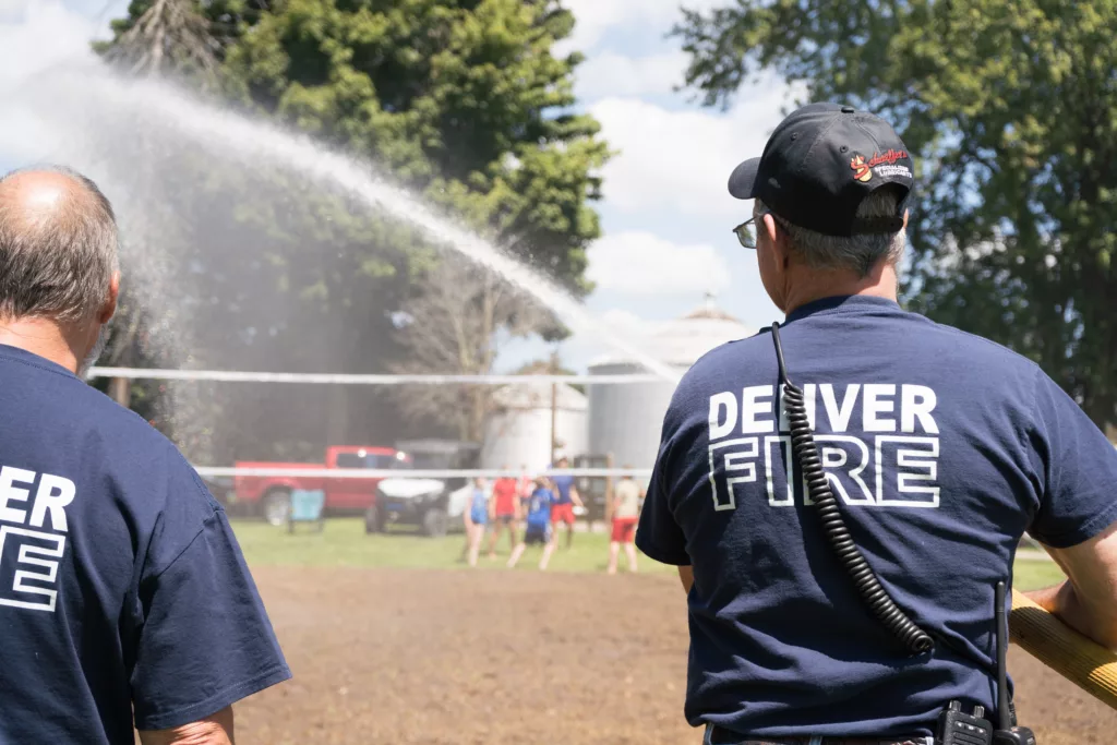 A firefighter stands in the left of a the frame and sprays a stream of water onto a dirt volleyball court.