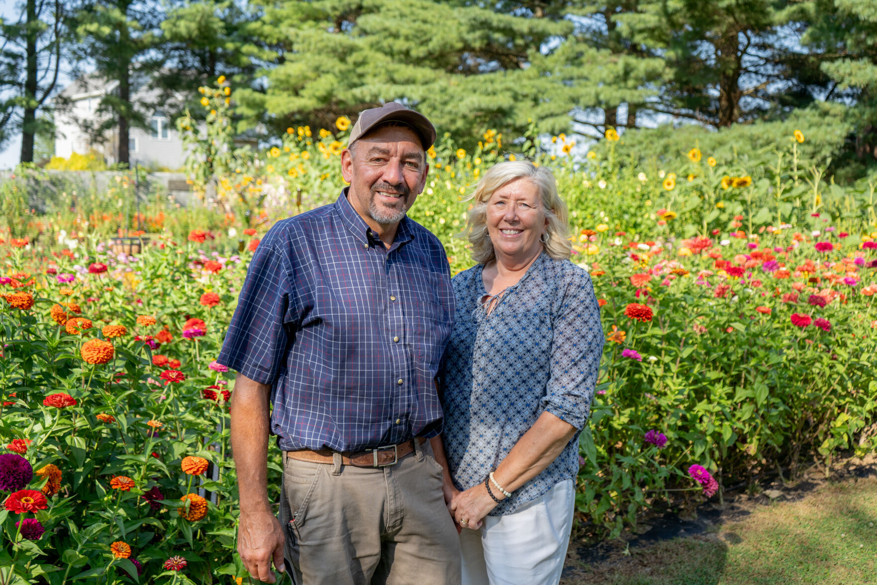 Michael Arthur, who is wearing a baseball cap, a blue shirt, and beige pants stands next to a Jayme arthur, who is wearing a blue blouse and blue jeans. They are posed in front of a wildflower patch with red and orange flowers.